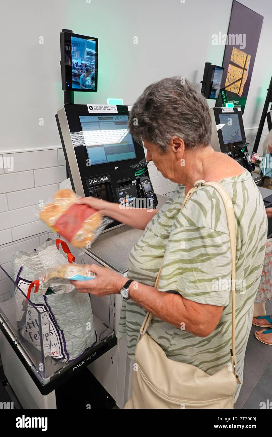 Scansione e pagamento dei prodotti alimentari interni M&S fino a una donna anziana che passa il cibo al bar scanner e schermo della fotocamera di sicurezza sul muro Inghilterra Regno Unito Foto Stock