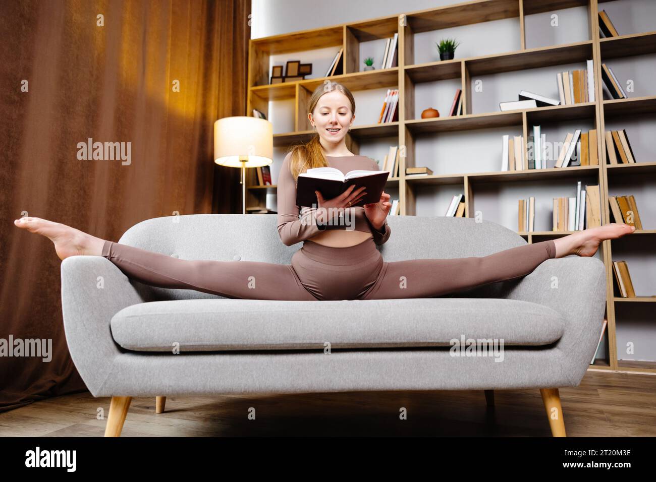 Giovane bella donna bionda in leggings marrone e top seduta su uno spago a casa sul divano. La ragazza sta leggendo un libro seduto sul divano in un Foto Stock