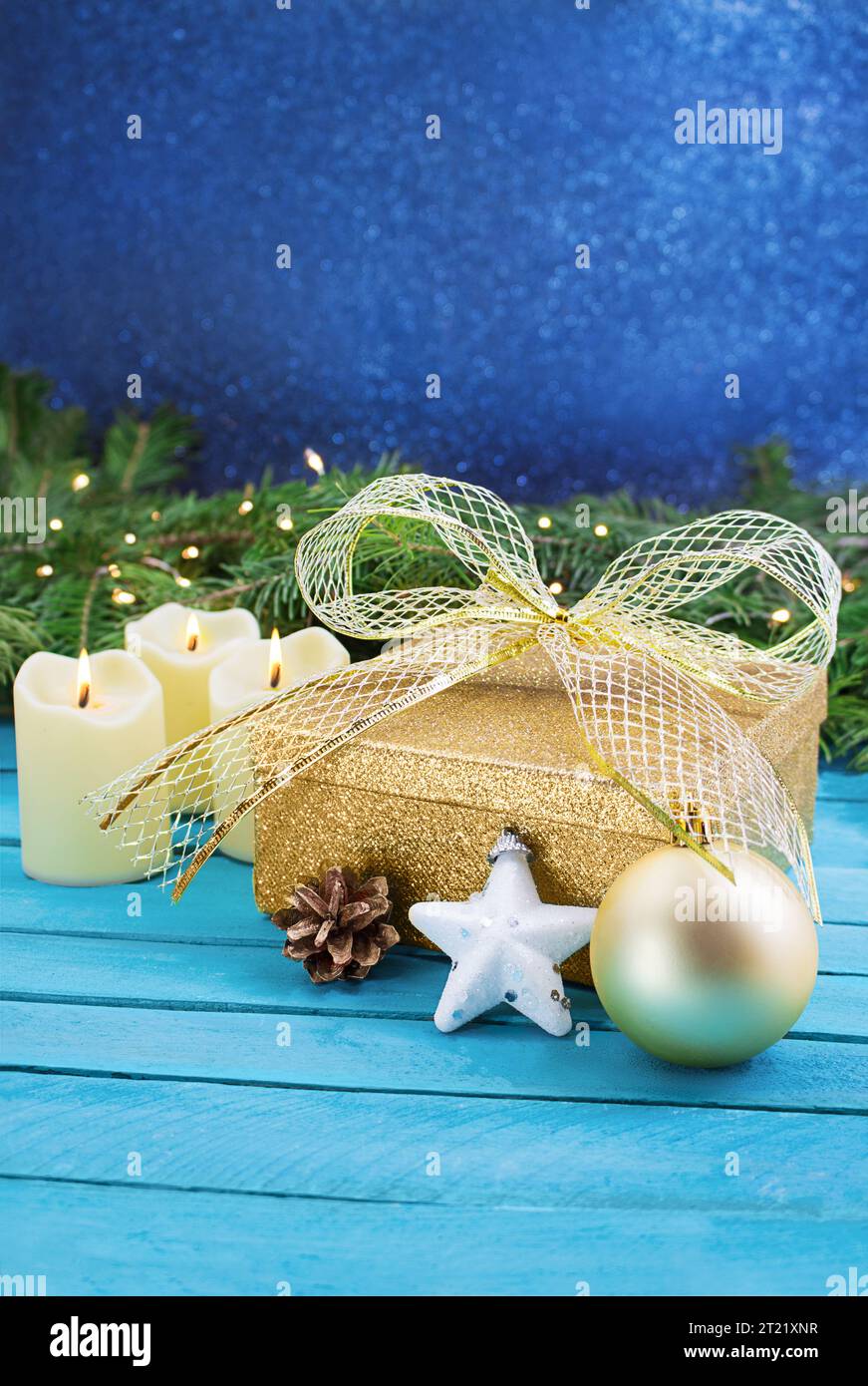 Confezione regalo giallo dorato con fiocco, candele bruciate, stella bianca e cono di pino su tavole blu dietro il ramo di abete con sfondo sfocato. Natale, New Ye Foto Stock