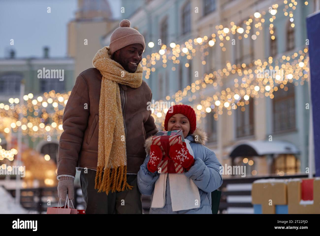 Riduci il ritratto della bambina nera che fa shopping natalizio con il padre e tiene le confezioni regalo all'aperto in inverno Foto Stock