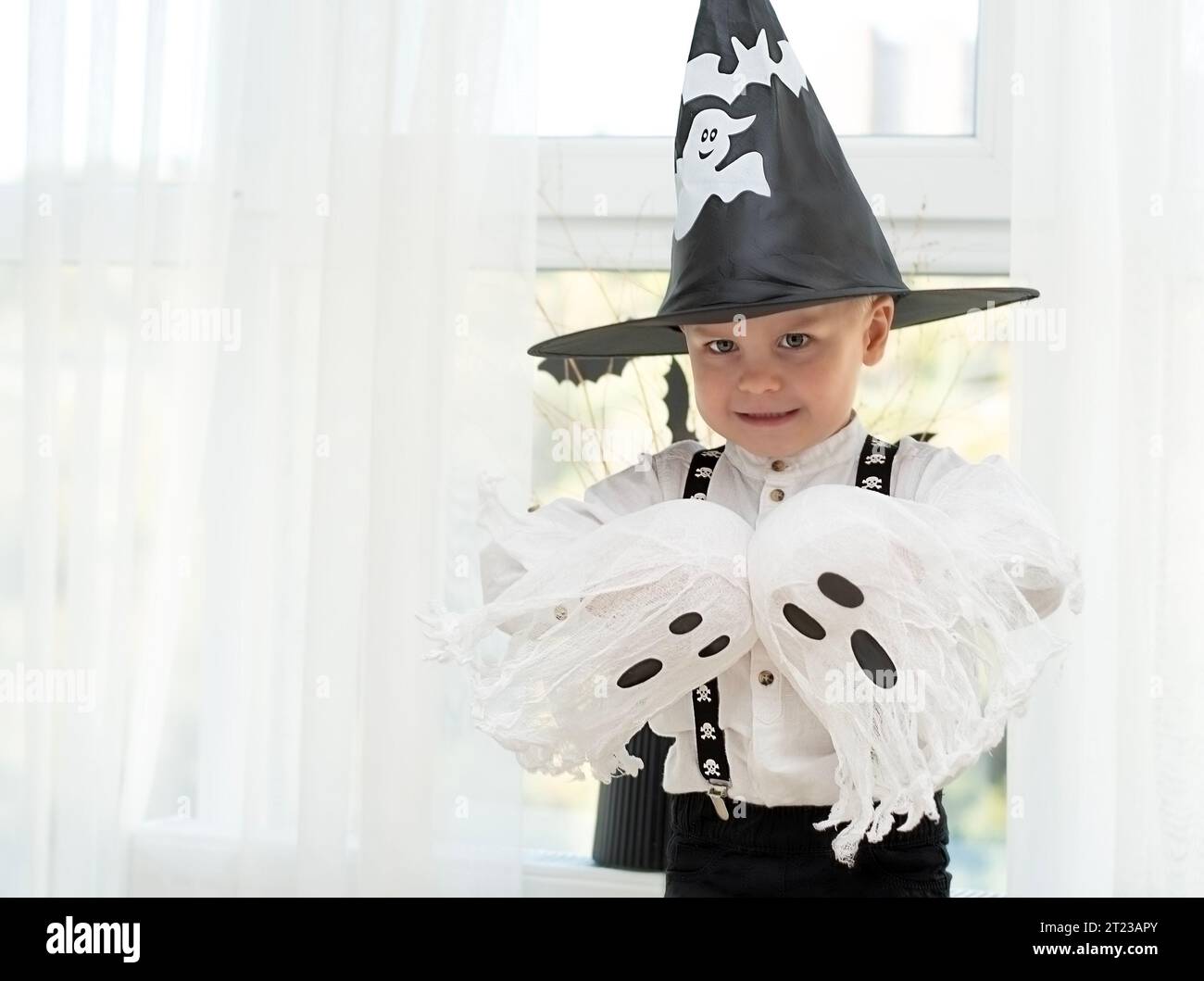 Concetto di Halloween. Un piccolo e bello ragazzo con il cappello di un mago con fantasmi bianchi gioca allegramente ed emotivamente all'interno di una casa sullo sfondo Foto Stock