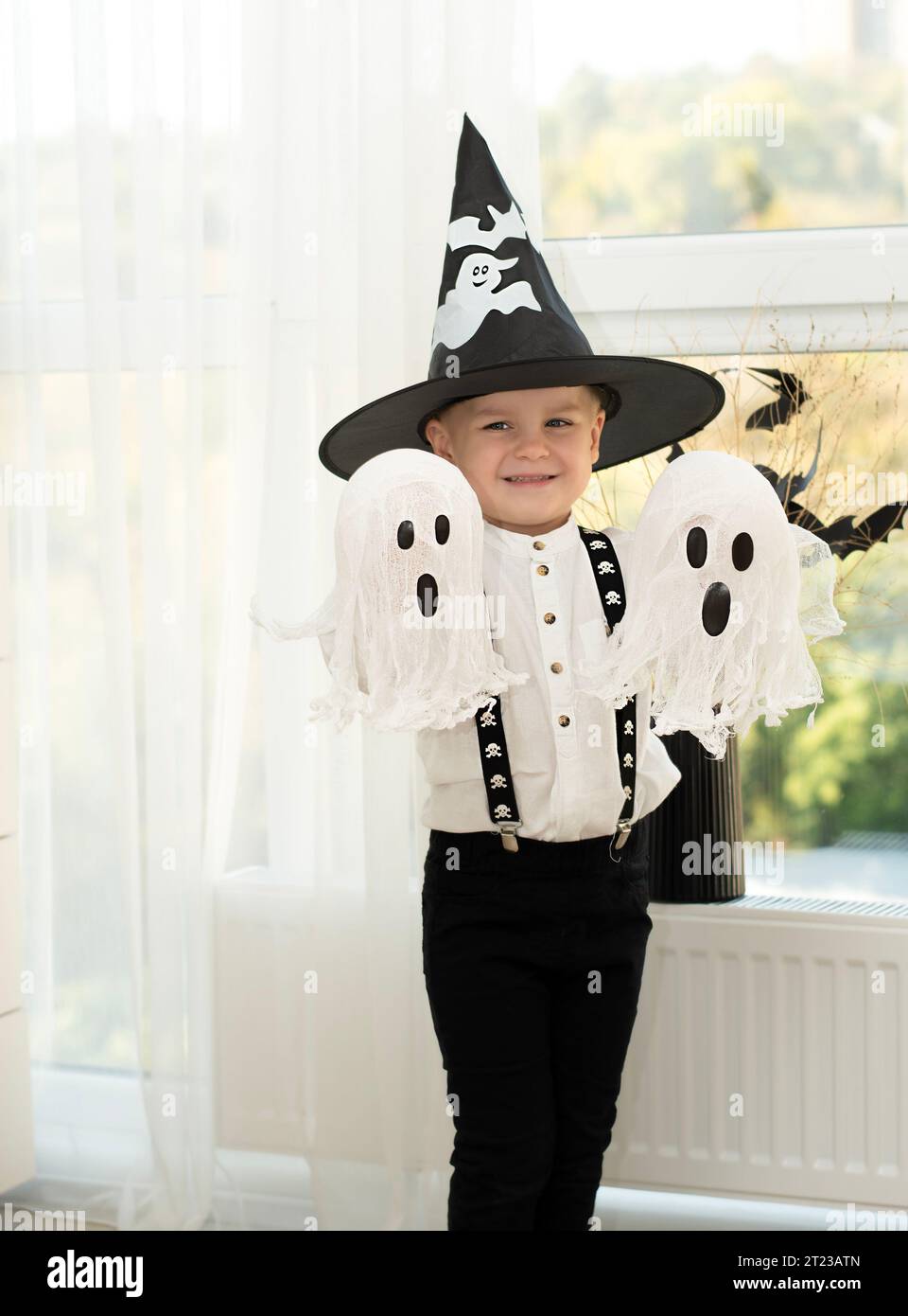 Concetto di Halloween. Un piccolo e bello ragazzo con il cappello di un mago con fantasmi bianchi gioca allegramente ed emotivamente all'interno di una casa sullo sfondo Foto Stock