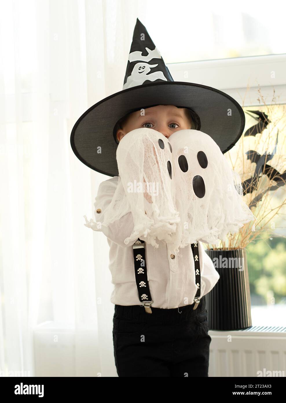 Concetto di Halloween. Un piccolo e bello ragazzo con il cappello di un mago con fantasmi bianchi gioca allegramente ed emotivamente all'interno di una casa sullo sfondo Foto Stock
