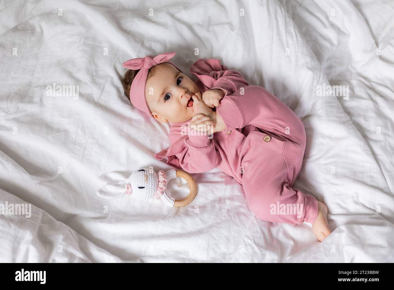 Il bambino in una tuta rosa con un sonaglino di legno ecologico è sdraiato a letto Foto Stock