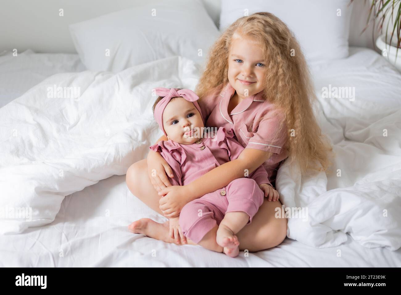 Graziosa ragazza riccia in pigiama rosa tiene un bambino tra le braccia mentre è seduto a letto Foto Stock