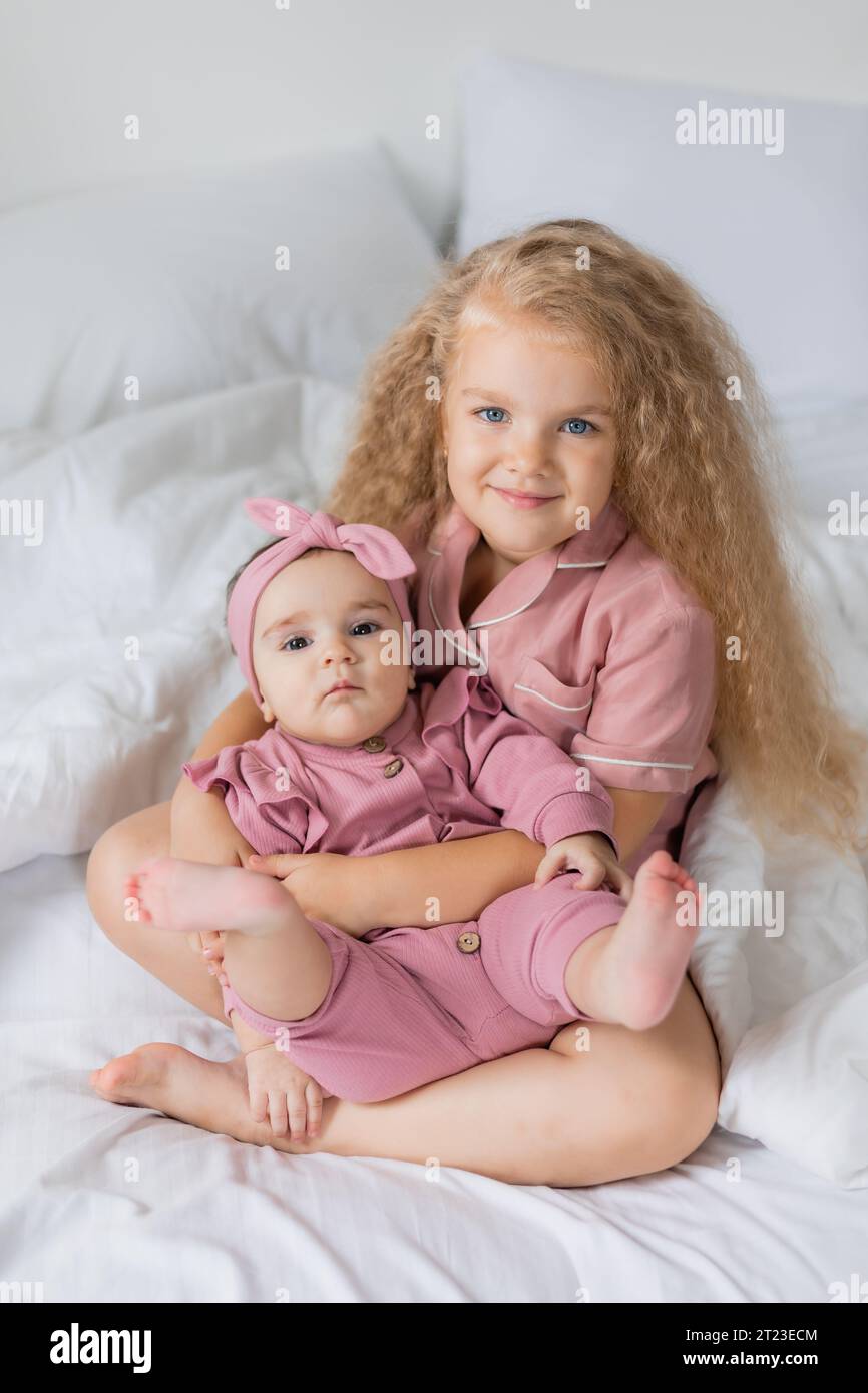 Graziosa ragazza riccia in pigiama rosa tiene un bambino tra le braccia mentre è seduto a letto Foto Stock