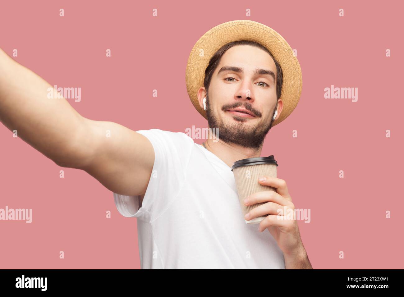 Ritratto di un uomo blogger barbuto positivo e sorridente in maglietta bianca e cappello con caffè e foto a vista. Riprese in studio per interni isolate su sfondo rosa. Foto Stock