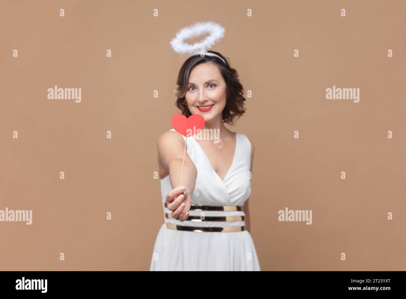 Ritratto di donna angelica con capelli ondulati e nimb sopra la testa, regalando cuori rossi, congratulandosi con San Valentino, indossando un abito bianco. Riprese in studio in interni isolate su sfondo marrone chiaro. Foto Stock