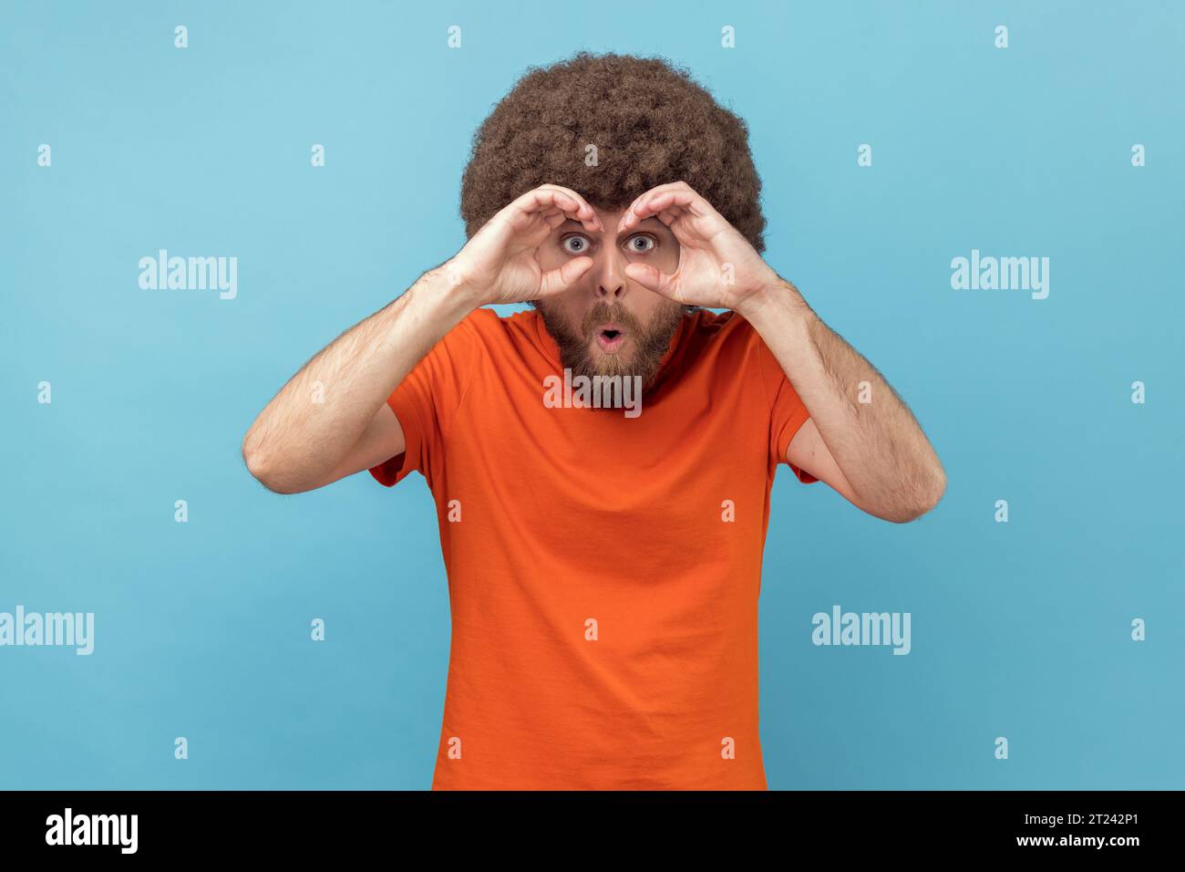 Ritratto di un uomo con acconciatura afro che indossa una T-shirt arancione che tiene i pugni vicino agli occhi immaginando un binocolo e guardando attraverso i buchi, spiando e divertendosi. Riprese in studio in interni isolate su sfondo blu. Foto Stock