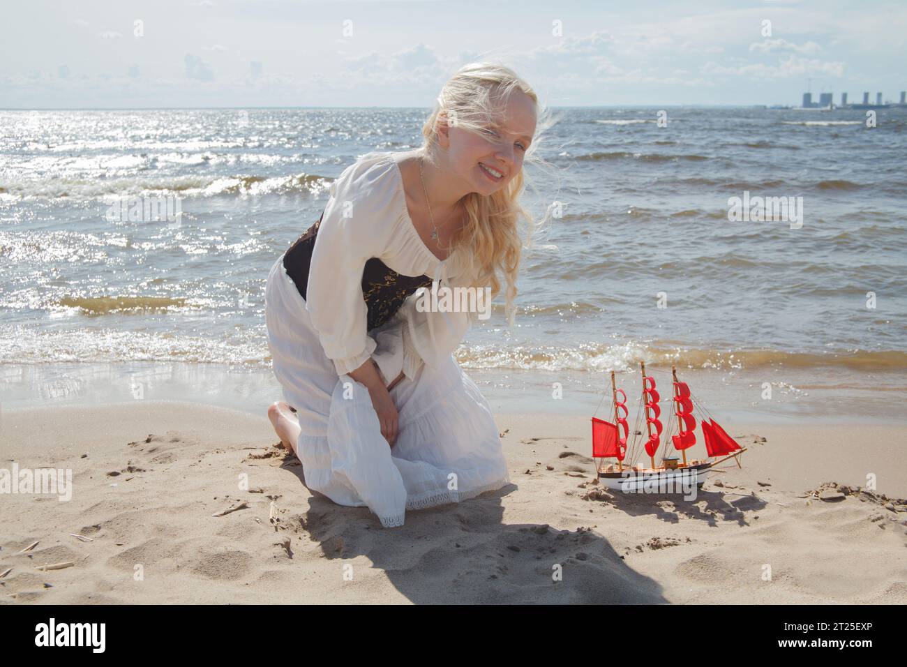Felice e attraente donna bionda con barca a vela rossa sulla spiaggia di mare. Concetto di vacanza romantica Foto Stock