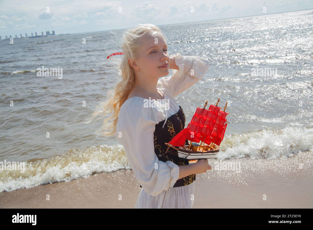 Giovane donna romantica e attraente che indossa un abito bianco d'epoca e regge una barca a vela rossa in piedi sulla spiaggia di mare Foto Stock