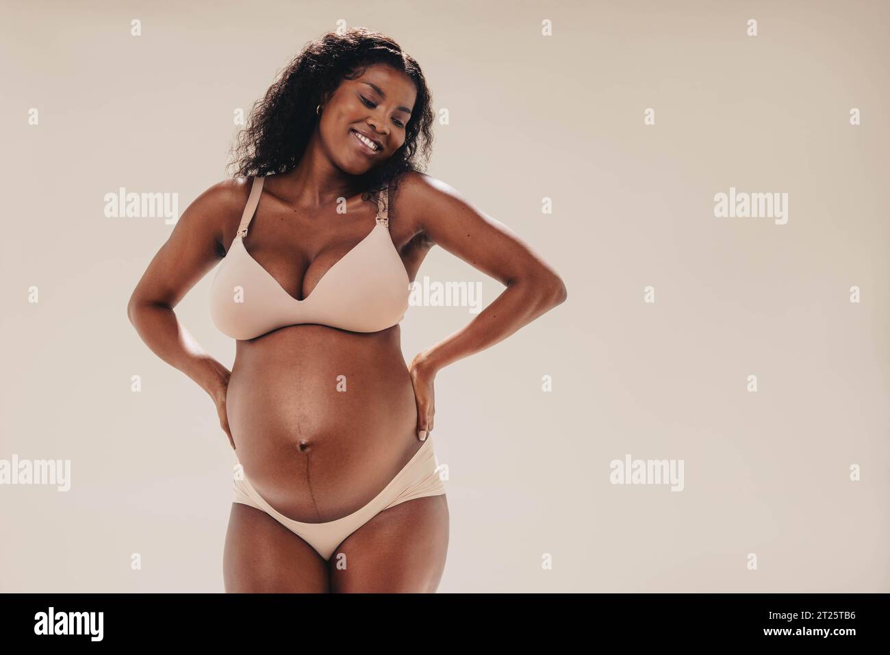 Una donna nera felice e sicura nel suo terzo trimestre si trova in uno studio, mostrando il suo bellissimo baby bump. Indossa lingerie, abbracciando la melanina Foto Stock