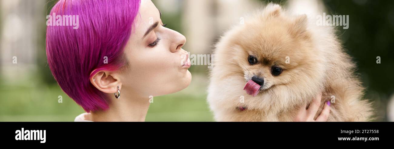 donna dai capelli viola che si fa strada sulle labbra e tiene in mano il divertente pomeranian spitz che si staglia sulla lingua e lo striscione Foto Stock