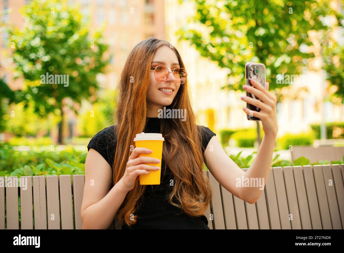 Sorridente ragazza divertente con occhiali da sole freschi utilizzando i social media con uno smartphone seduto su una panchina in città, una tazza di caffè di carta usa e getta Foto Stock