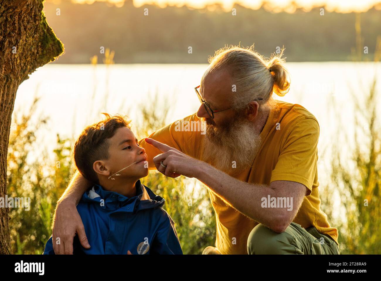 Un momento commovente come padre e figlio seduto nell'erba vicino a un lago forestale al tramonto, condividendo un legame speciale nella bellezza serena della natura. Padre e figlio Bonding a Sunset Lake. Foto di alta qualità Foto Stock