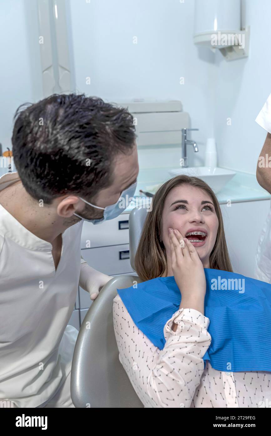 Una ragazza dall'aspetto preoccupata sulla sedia del dentista davanti al dottore si lamenta per il suo dolore gengivale e per aver tenuto la mano sulla guancia. Foto Stock