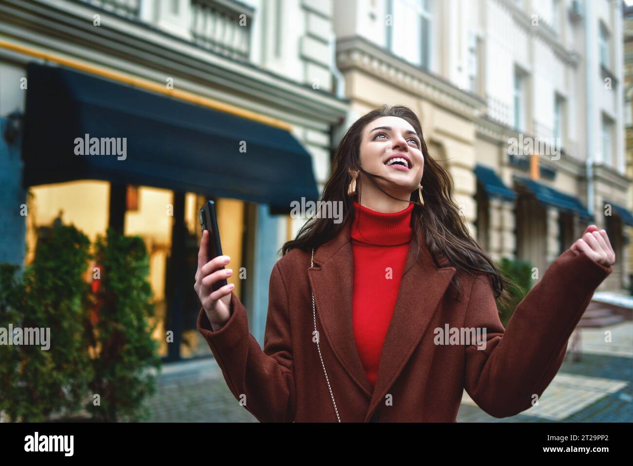 Eccitata donna alla moda che festeggia le buone notizie tenendo il telefono in strada la sera Foto Stock