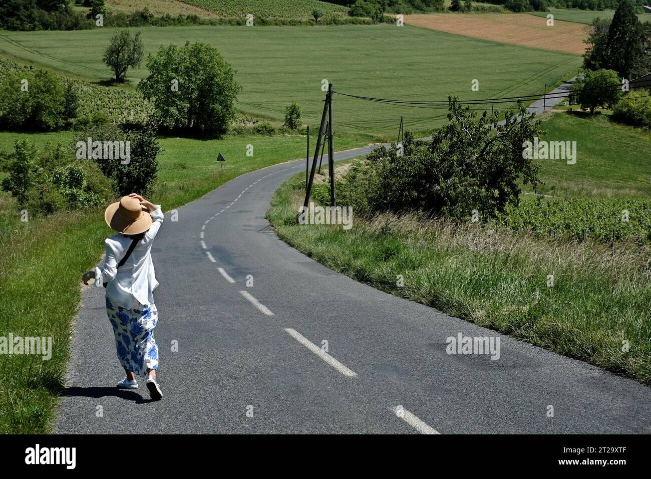 Una donna che indossa un cappello da sole che cammina lungo le tortuose strade di campagna francesi, attraverso vigneti e campi di grano Foto Stock