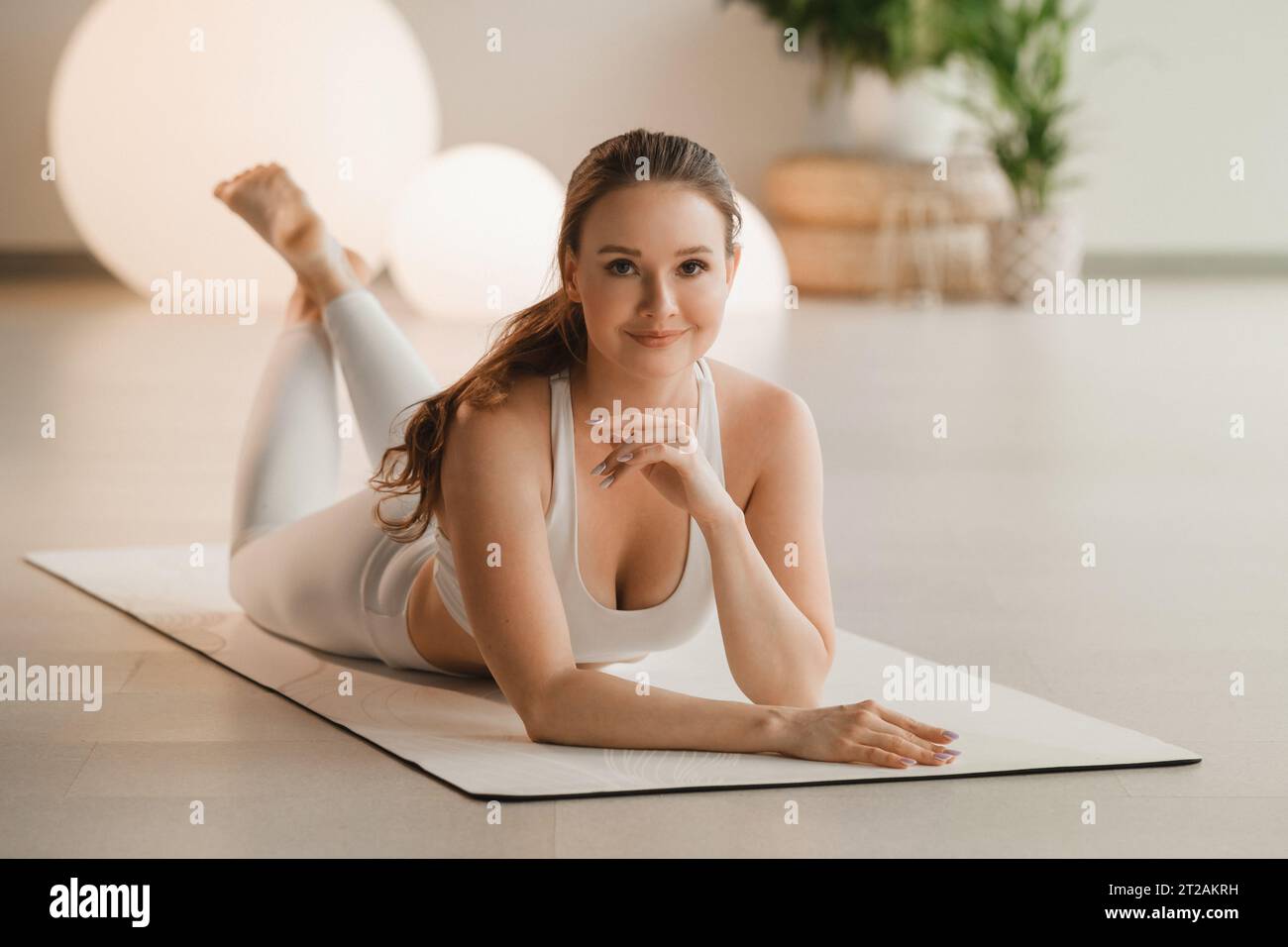 Ritratto di una ragazza in abiti bianchi sdraiata su un tappetino prima di fare yoga in interni. Foto Stock
