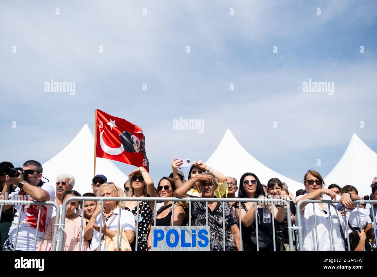 Smirne, Turchia, 9 settembre 2023: Una folla si trova dietro una barricata della polizia, tenendo alte le bandiere turche mentre osservano con attenzione le cerimonie che segnano i Foto Stock