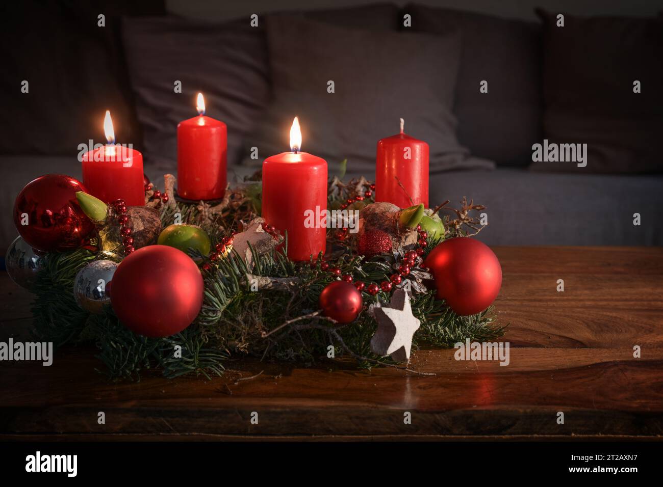 Corona dell'avvento con tre candele rosse brucianti e decorazioni natalizie su un tavolo di legno davanti al divano, decorazioni per la casa festiva per la terza domenica Foto Stock
