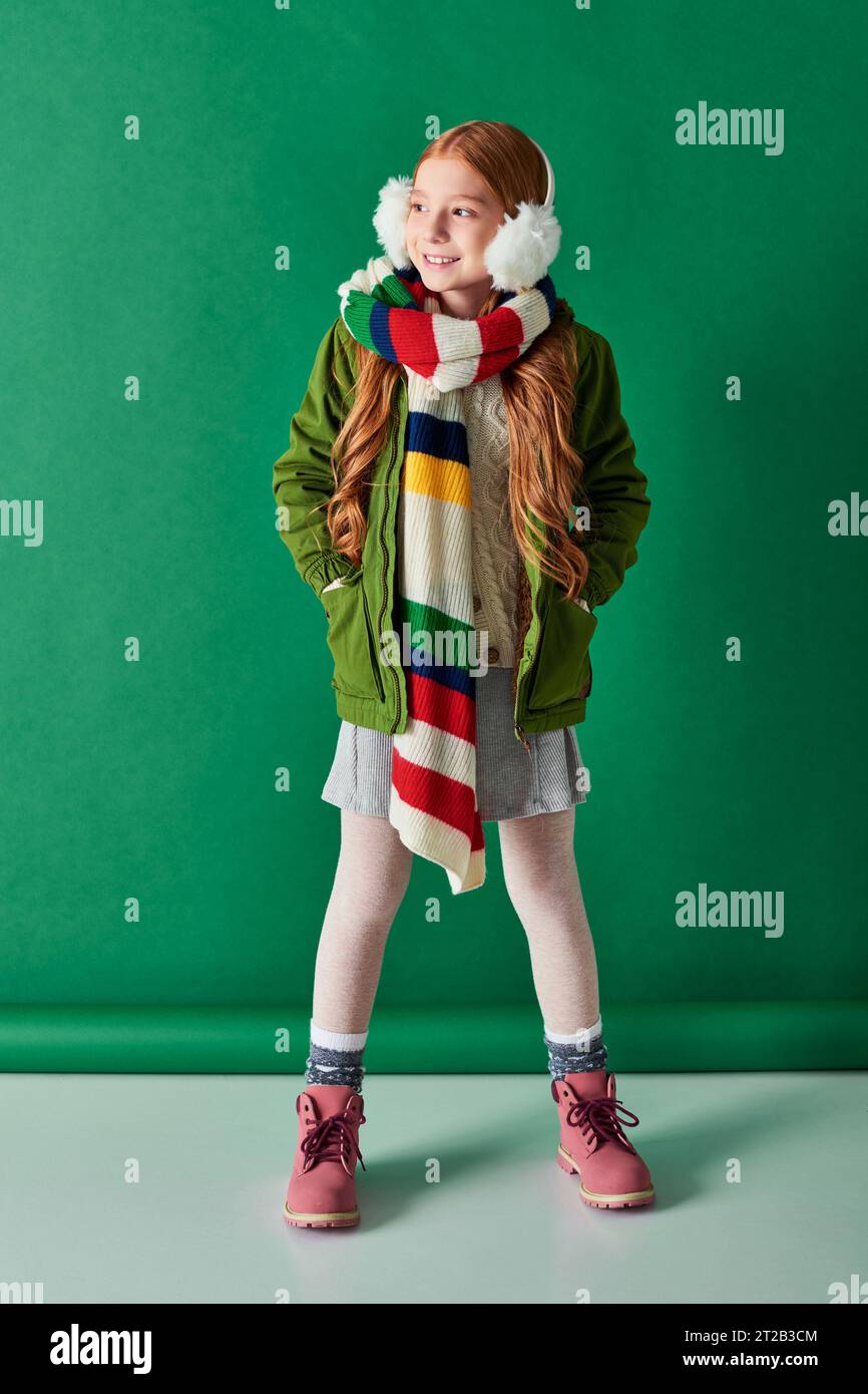 a tutta lunghezza, allegra ragazza con paraorecchie, sciarpa a righe e outfit invernale su sfondo turchese Foto Stock