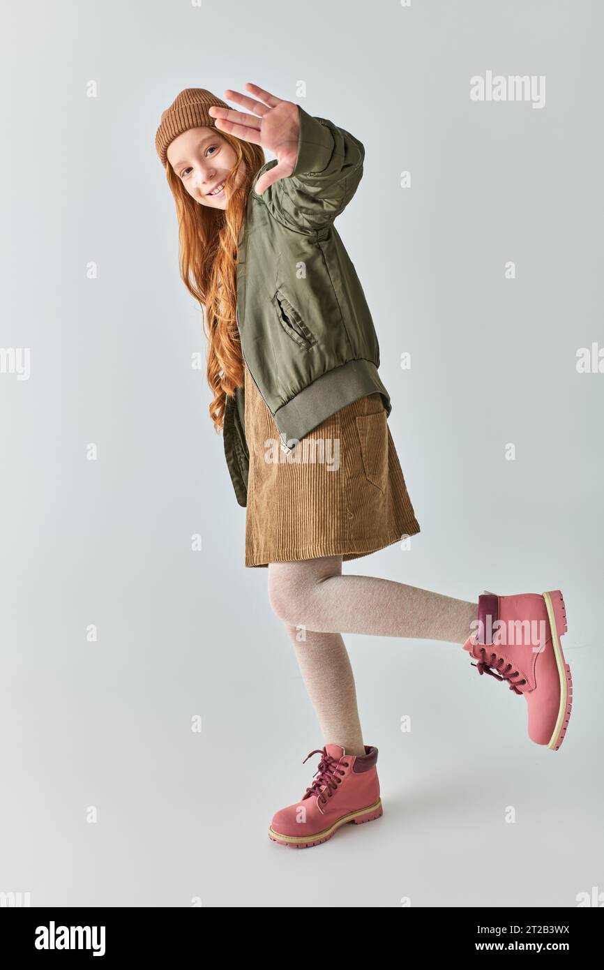 un'allegra ragazza che indossa un outfit invernale e un cappello a maglia che posa con la mano allungata sul grigio Foto Stock