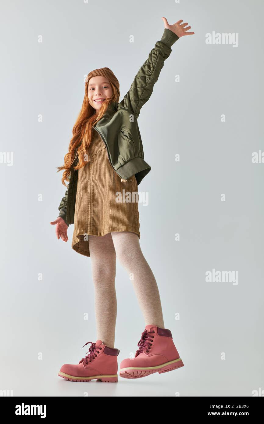 ragazza allegra con un elegante outfit invernale con stivali e cappello che sorridono e sventolano la mano su sfondo grigio Foto Stock