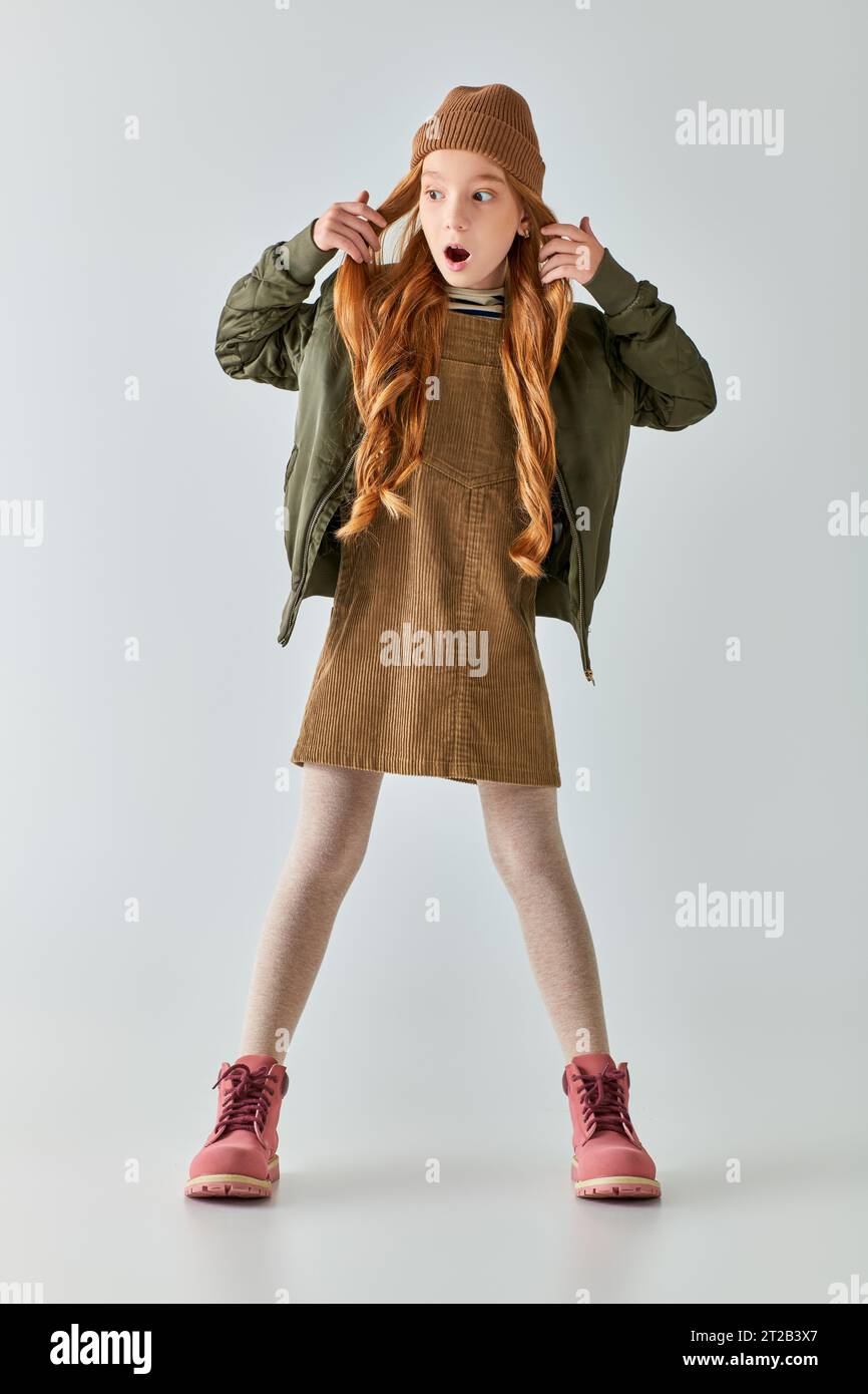 sbalordita preadolescente con i capelli lunghi in posa con un elegante abito invernale e un cappello su sfondo grigio Foto Stock