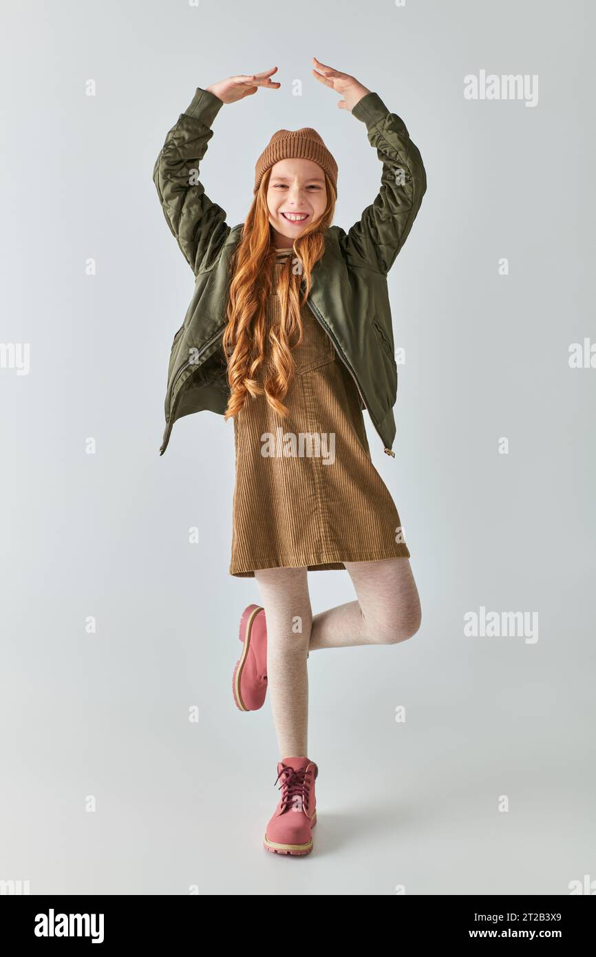 ragazza allegra in outfit invernale con stivali e cappello sorridenti e in posa da ballerina su sfondo grigio Foto Stock