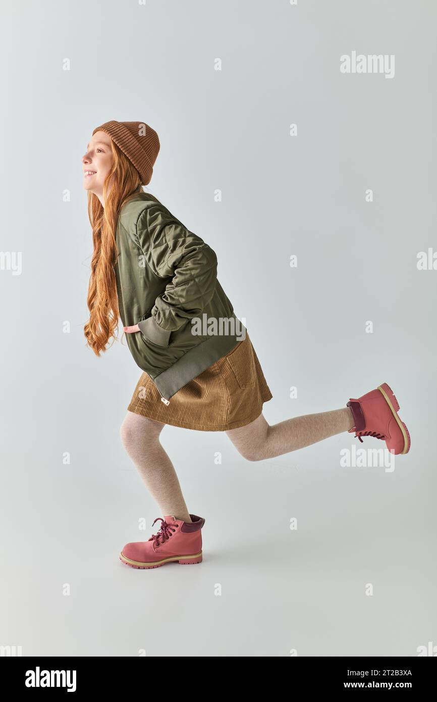 tutta la lunghezza della giocosa preadolescente in outfit invernale con stivali e cappello che corrono su sfondo grigio Foto Stock