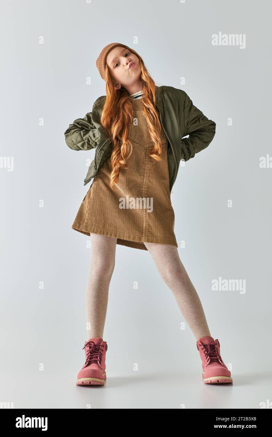 ragazza scontenta con un elegante outfit invernale e un cappello che riempie le guance e sta in piedi con le mani sui fianchi Foto Stock