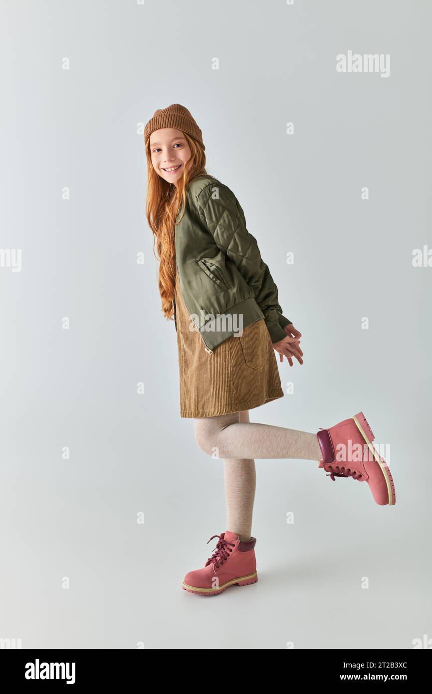 un'allegra ragazza preadolescente in completo invernale con stivali e cappello che guardano la fotocamera in grigio Foto Stock