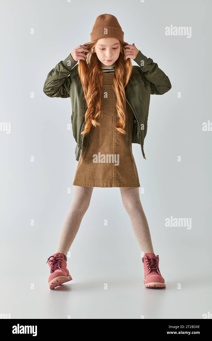 graziosa preadolescente con capelli lunghi in posa con un elegante abito invernale e un cappello a maglia su sfondo grigio Foto Stock