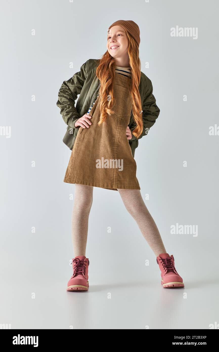 ragazza allegra con un elegante outfit invernale e un cappello sorridente e in piedi con le mani sui fianchi di colore grigio Foto Stock