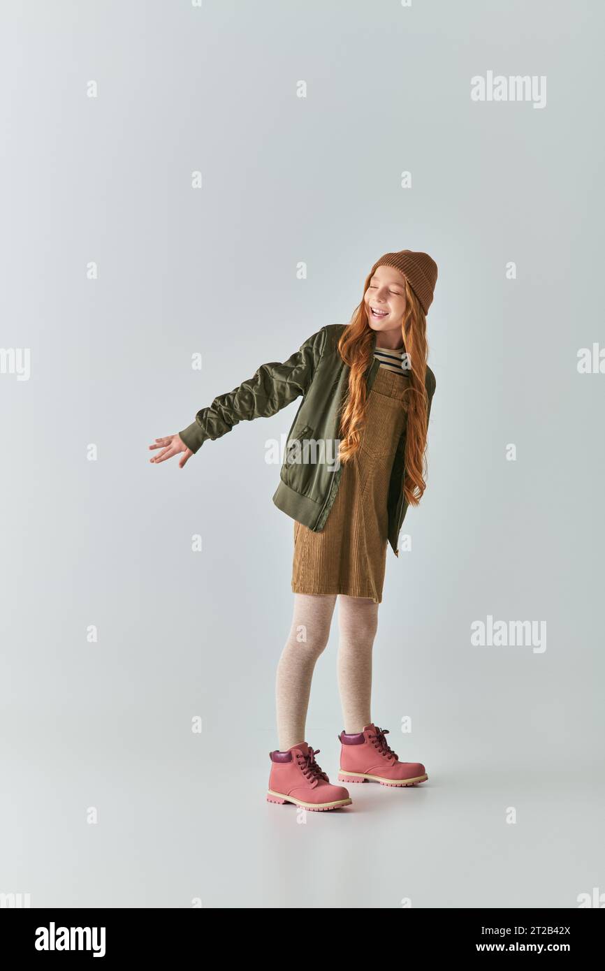 un'allegra ragazza che indossa un outfit invernale e un cappello a maglia che posa con le mani allungate sul grigio Foto Stock