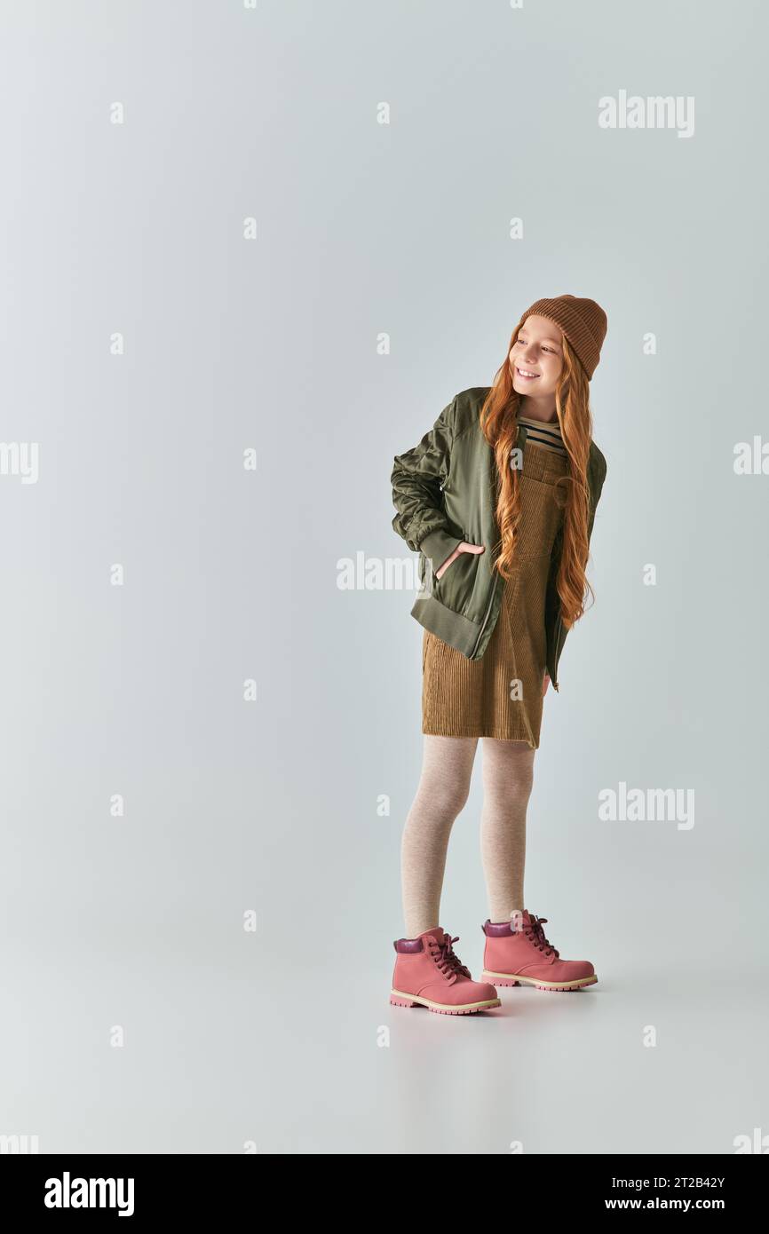 happy girl completa in outfit invernale e cappello a maglia in posa con la mano in tasca sul grigio Foto Stock