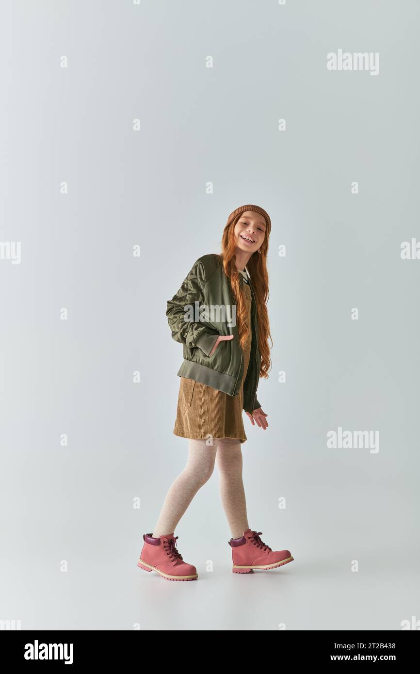un'allegra ragazza che indossa un outfit invernale e un cappello a maglia in posa con la mano in tasca sul grigio Foto Stock