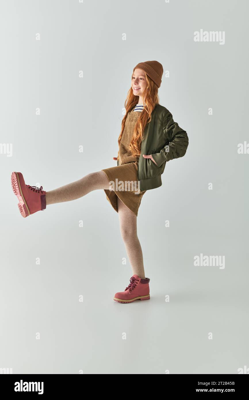 moda per bambini, ragazza gioiosa con capelli lunghi e cappello invernale sulla testa in piedi in abito e giacca Foto Stock