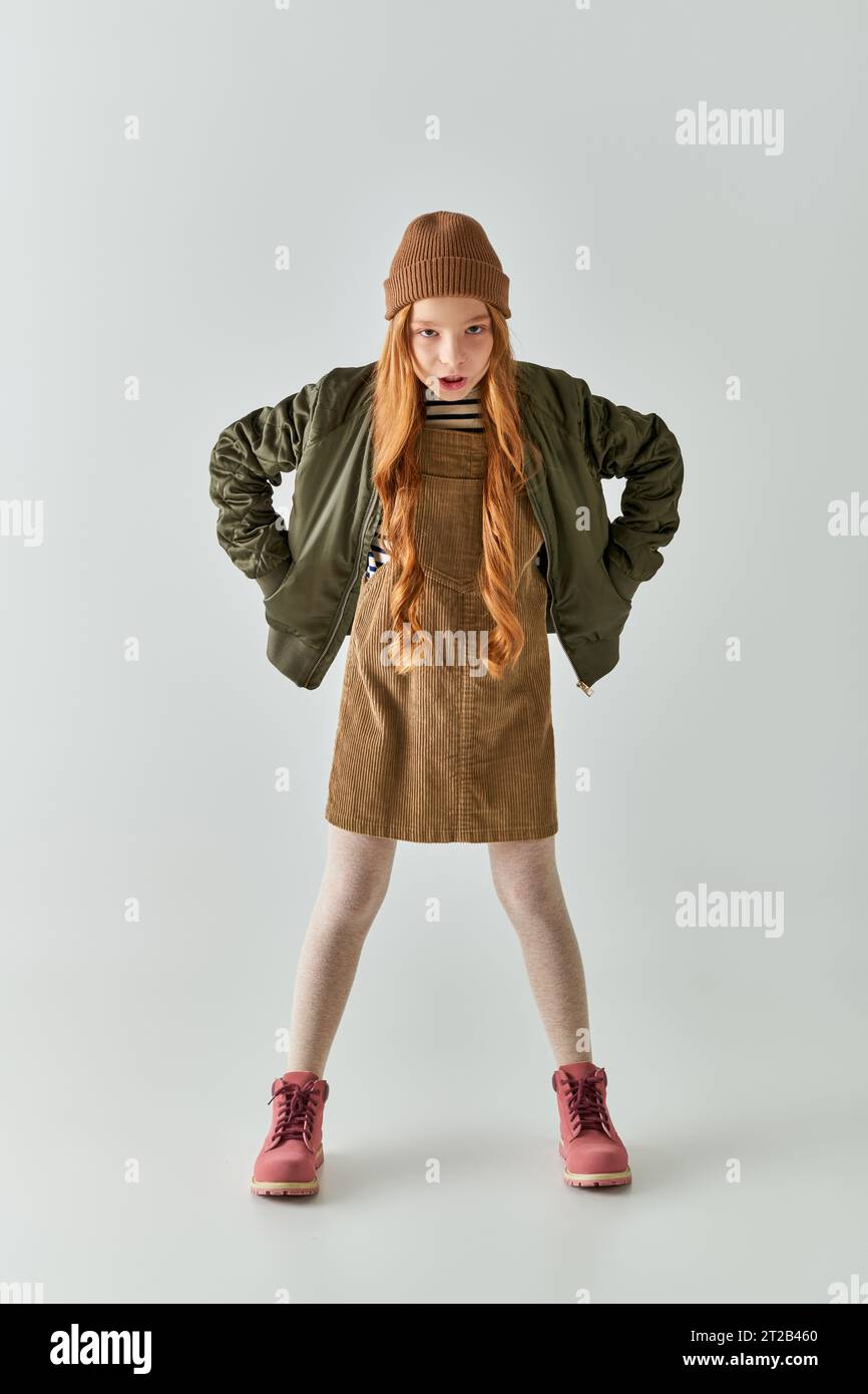 ragazza arrabbiata con capelli lunghi e cappello sulla testa in piedi in abito con giacca invernale su sfondo grigio Foto Stock