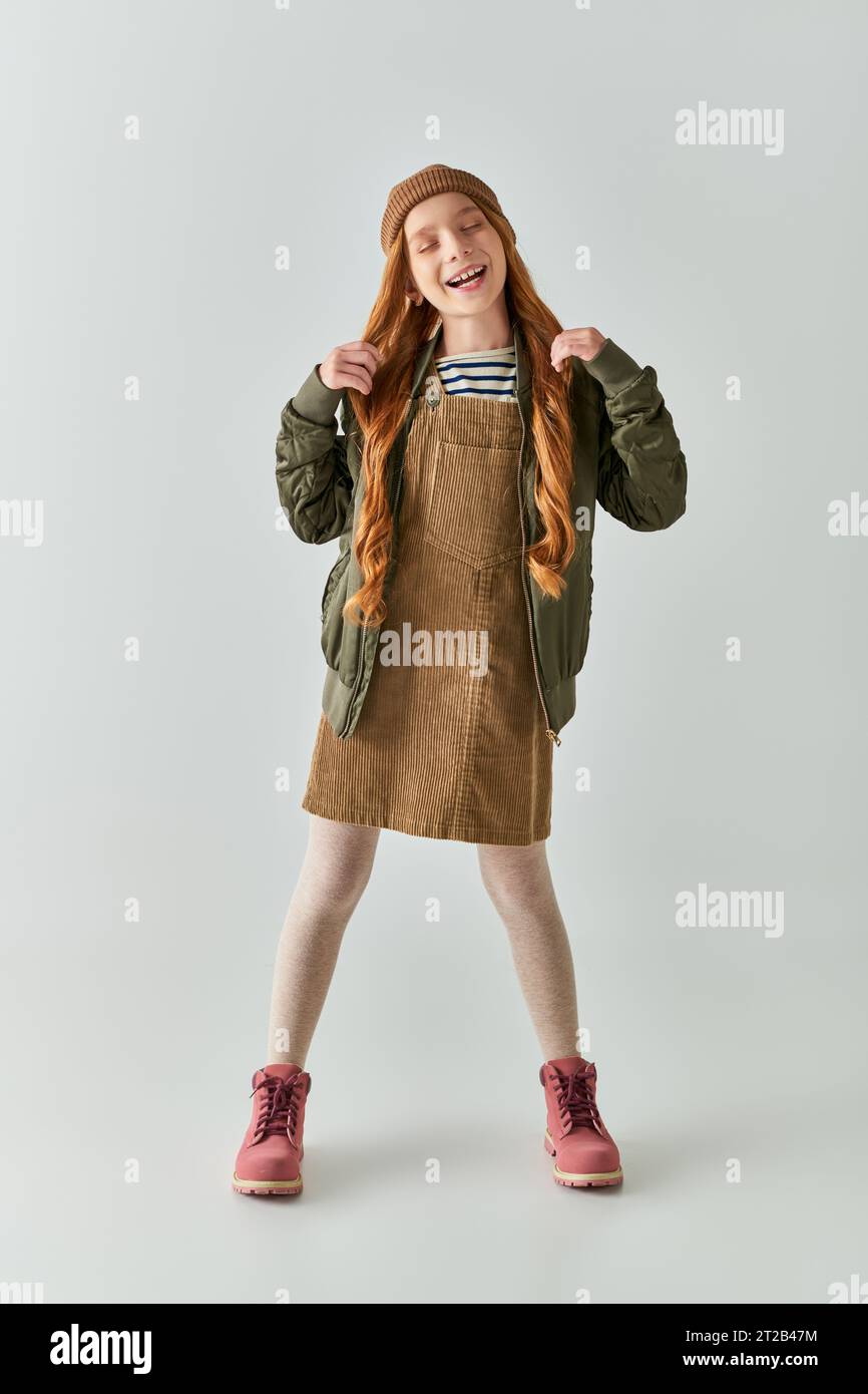 ragazza con capelli lunghi e cappello sulla testa in piedi in abito con giacca invernale su sfondo grigio Foto Stock