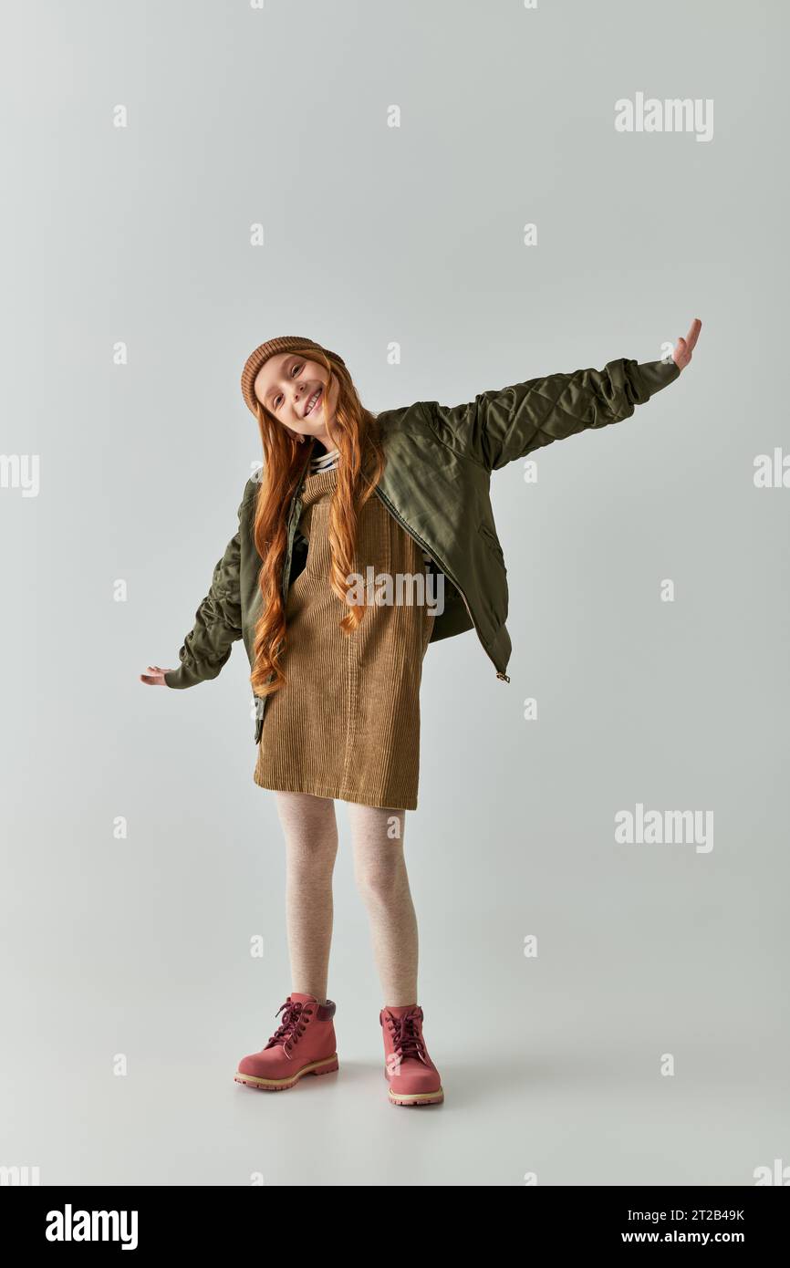 felice ragazza preadolescente con capelli lunghi in piedi con mani allungate su sfondo grigio, stile più avvolgente Foto Stock