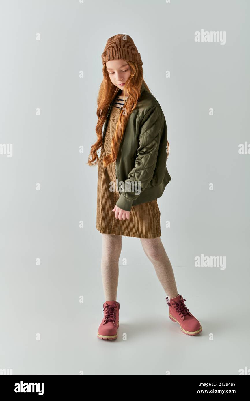 graziosa ragazza preadolescente con capelli lunghi in piedi in cappello, abito, giacca invernale e stivali su sfondo grigio Foto Stock