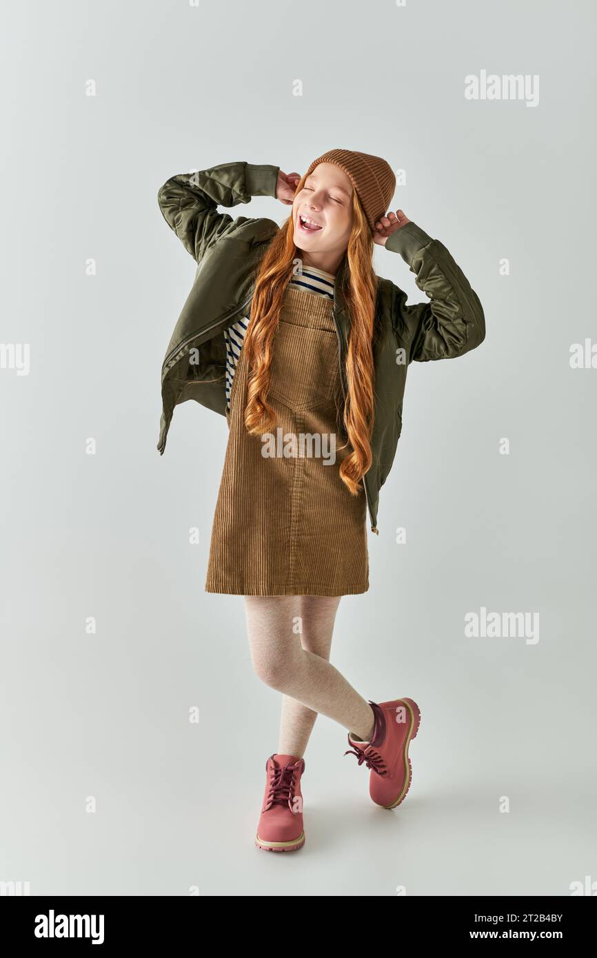 ragazza gioiosa con i capelli lunghi che indossa un cappello invernale e indossa un abito con giacca su sfondo grigio Foto Stock