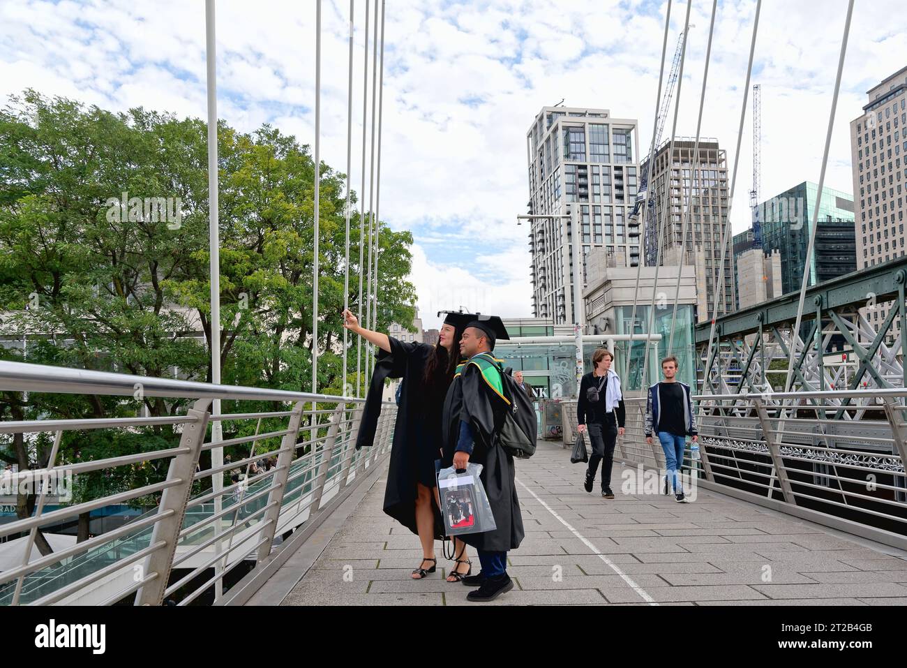 Una giovane coppia vestita di abito e mortaio si prende un selfie sul passaggio pedonale di South Bank, avendo appena partecipato a una cerimonia di laurea, Waterloo London. Foto Stock