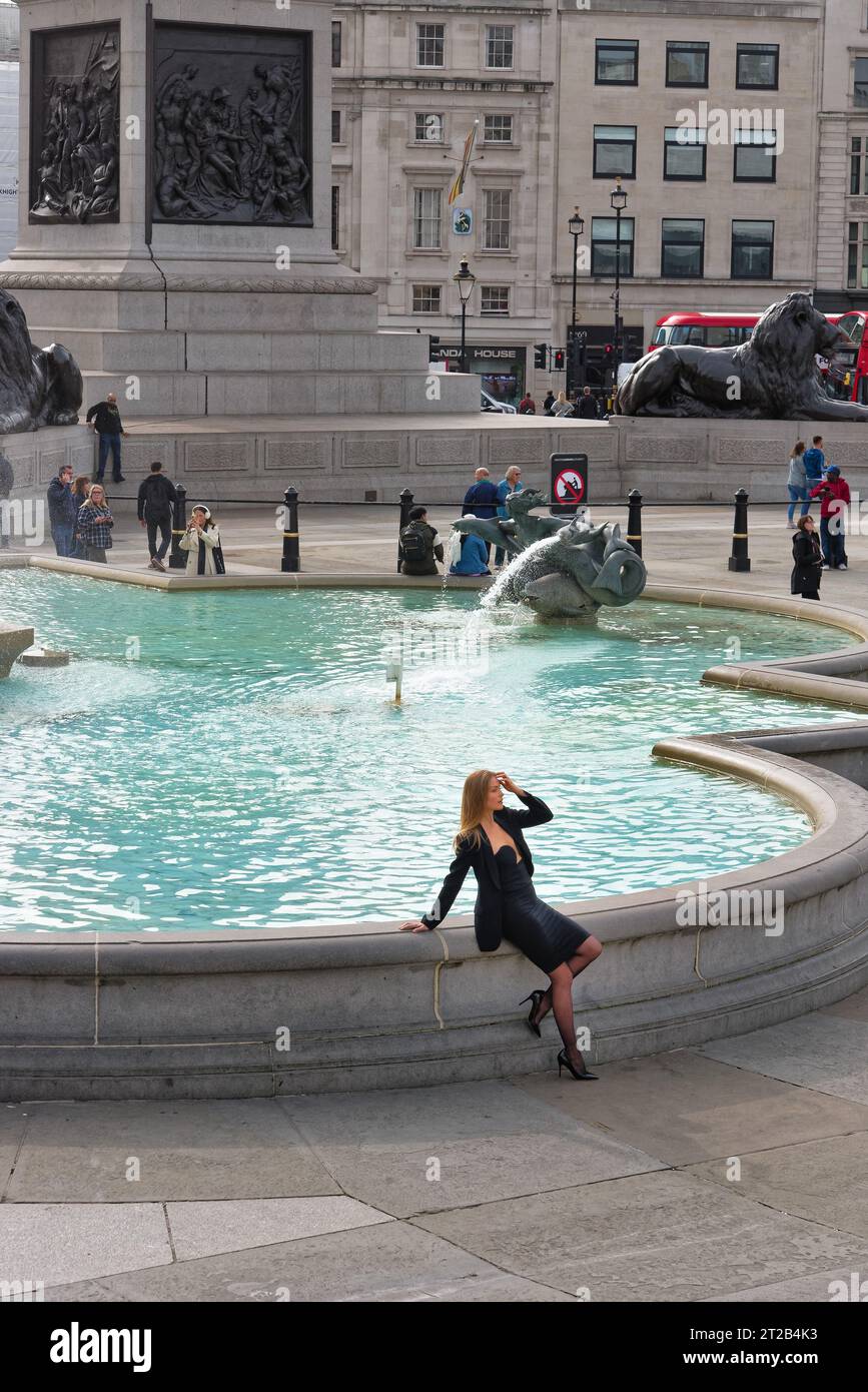 Un'attraente giovane donna europea bianca con un vestito nero che posa per le fotografie di una delle fontane di Trafalgar Square, nel centro di Londra, Inghilterra, Regno Unito Foto Stock