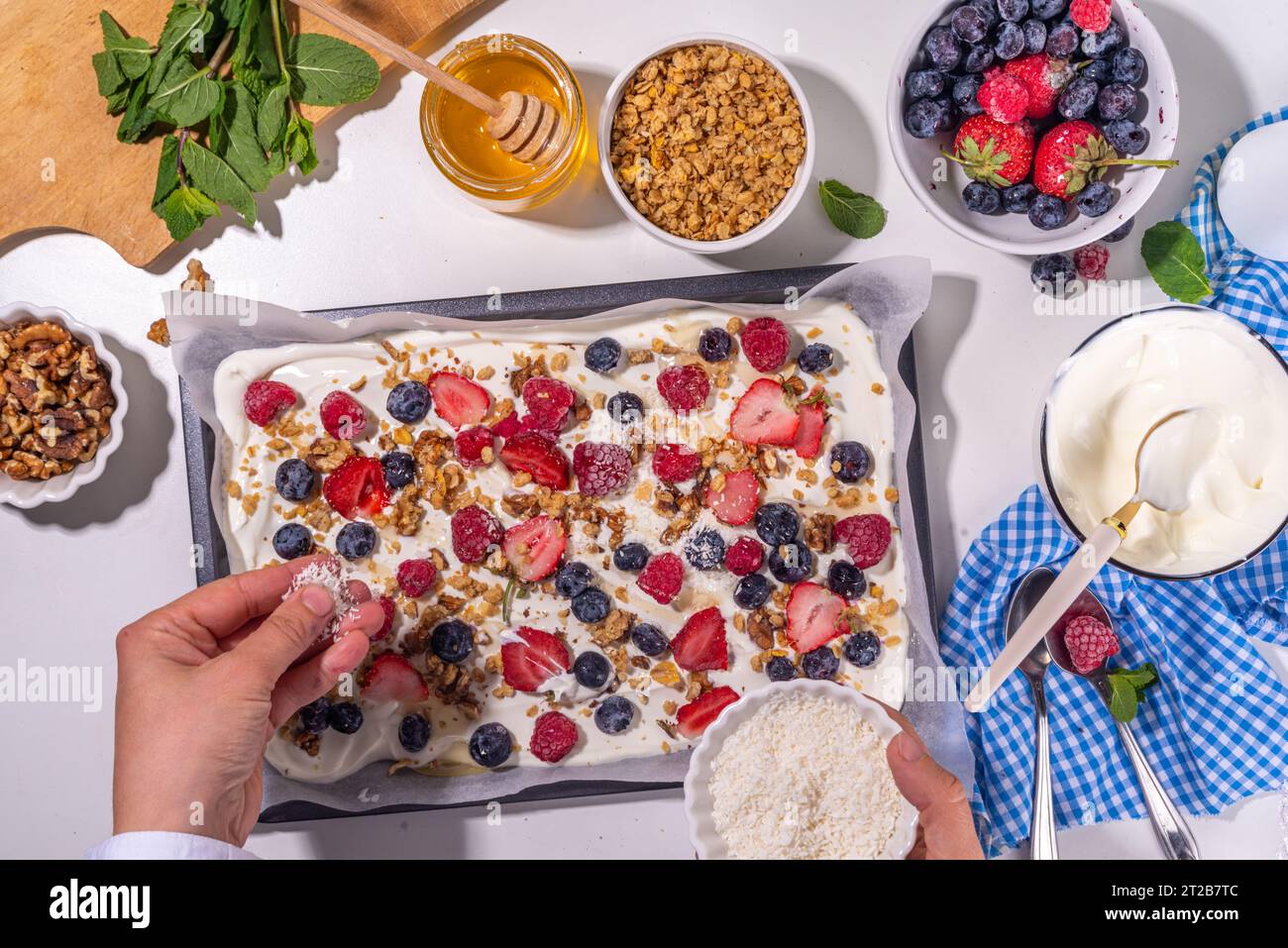 Preparazione della cottura con sfondo di corteccia di yogurt congelato con yogurt greco, bacche fresche, muesli, miele, salsa al cioccolato, con vassoio da forno deco. Vista dall'alto della donna Foto Stock