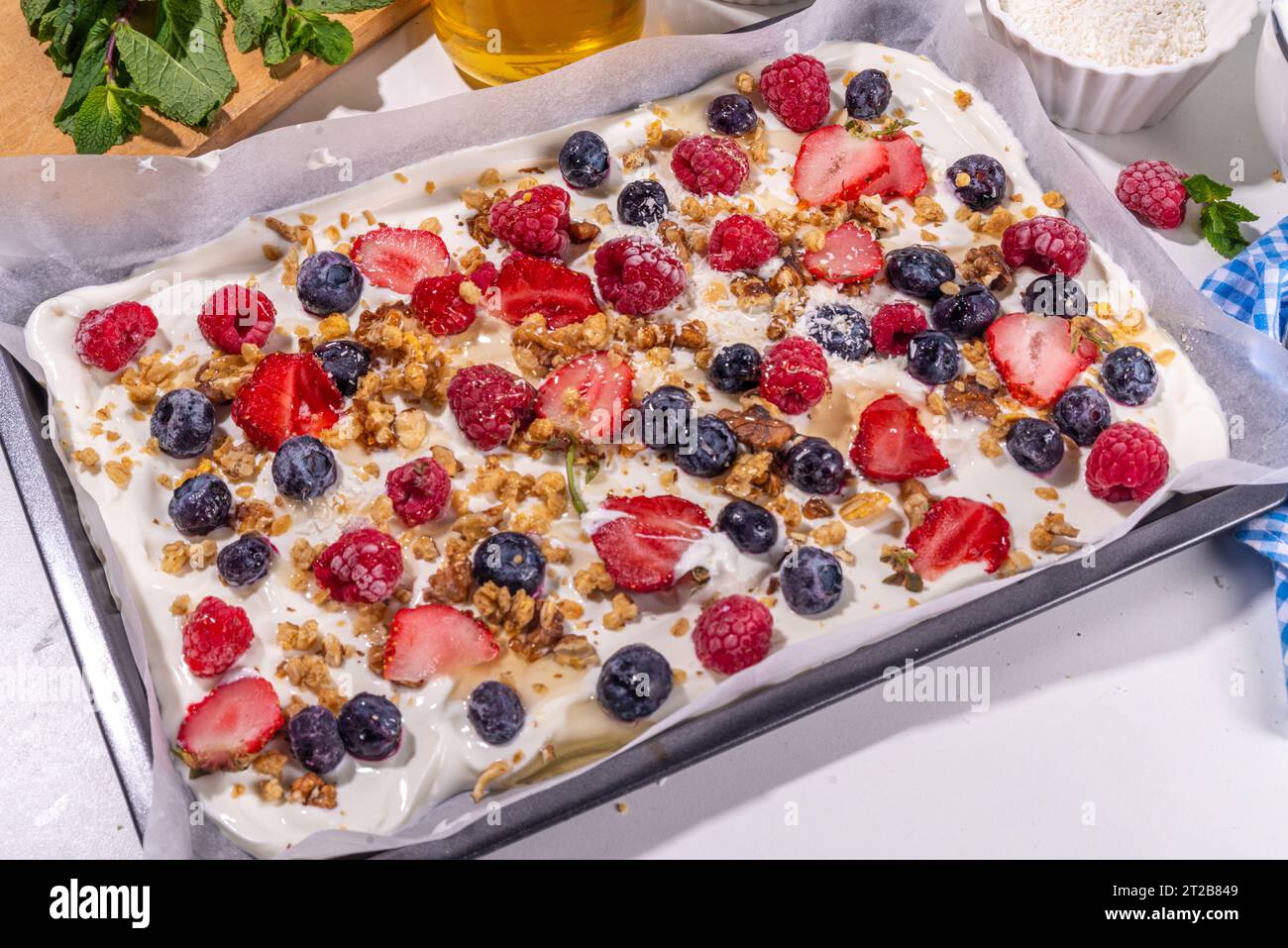 Preparazione della cottura con sfondo di corteccia di yogurt congelato con yogurt greco, bacche fresche, muesli, miele, salsa al cioccolato, con vassoio da forno deco. Vista dall'alto della donna Foto Stock