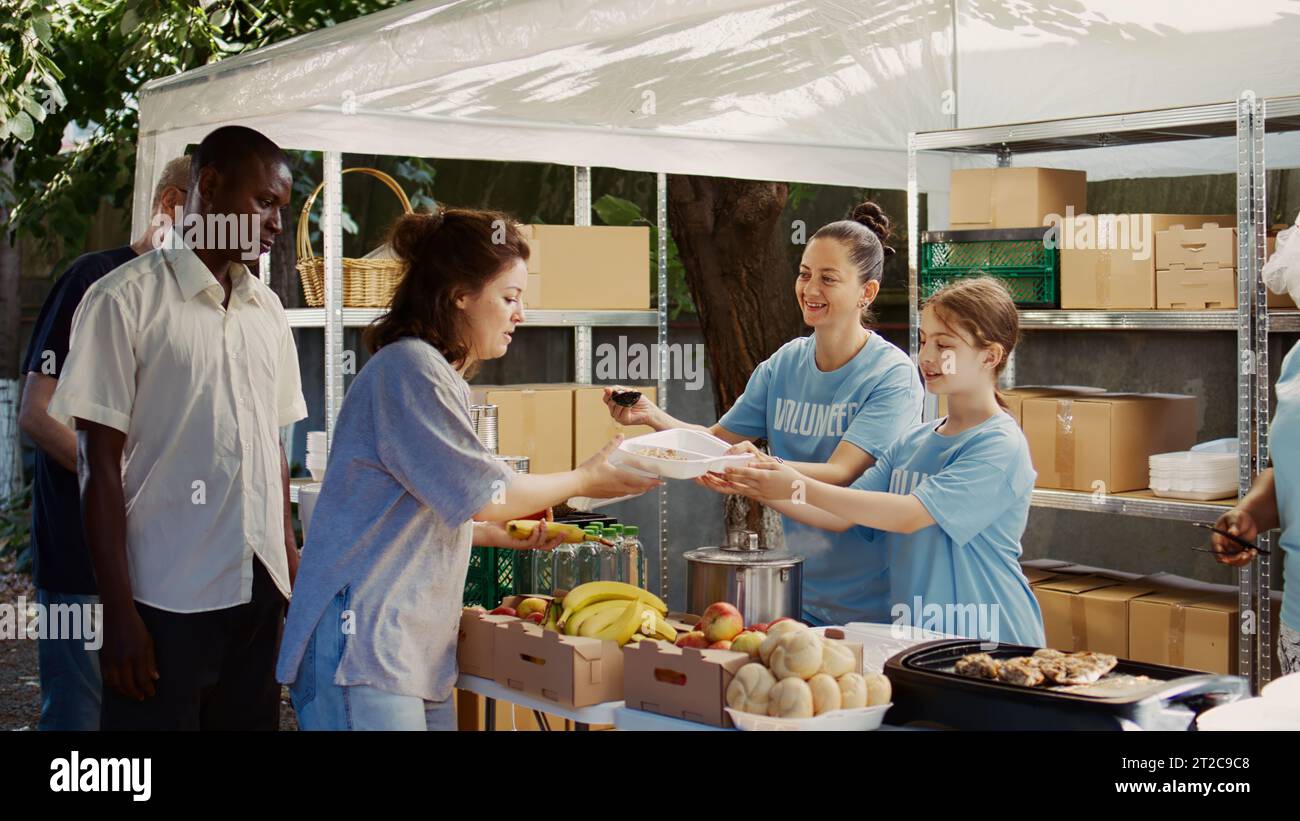 La ragazza caucasica confeziona cibo gratuito da distribuire ai più svantaggiati con l'assistenza di altri operatori di beneficenza. Volontarie che servono pasti caldi ai senzatetto e affamati presso la banca di cibo all'aperto. Foto Stock
