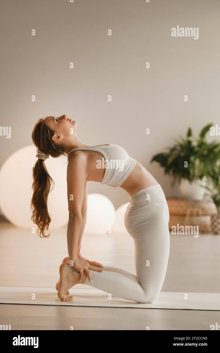 Una ragazza in abiti bianchi fa yoga su un tappetino al chiuso. Foto Stock